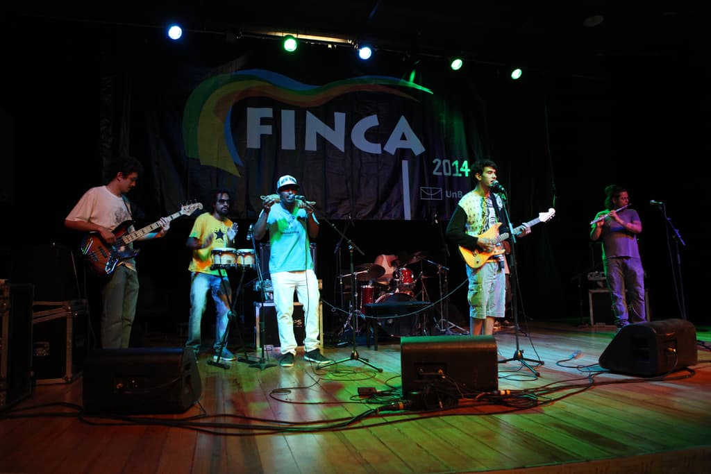 Fünf Personen auf der Bühne stehend und Musik performend mit einem Banner und Vorhang mit der Aufschrift "FINCA 2014" im Hintergrund, fünf Lampen an der Decke und Kabel und ein Musikkasten auf dem Boden.
