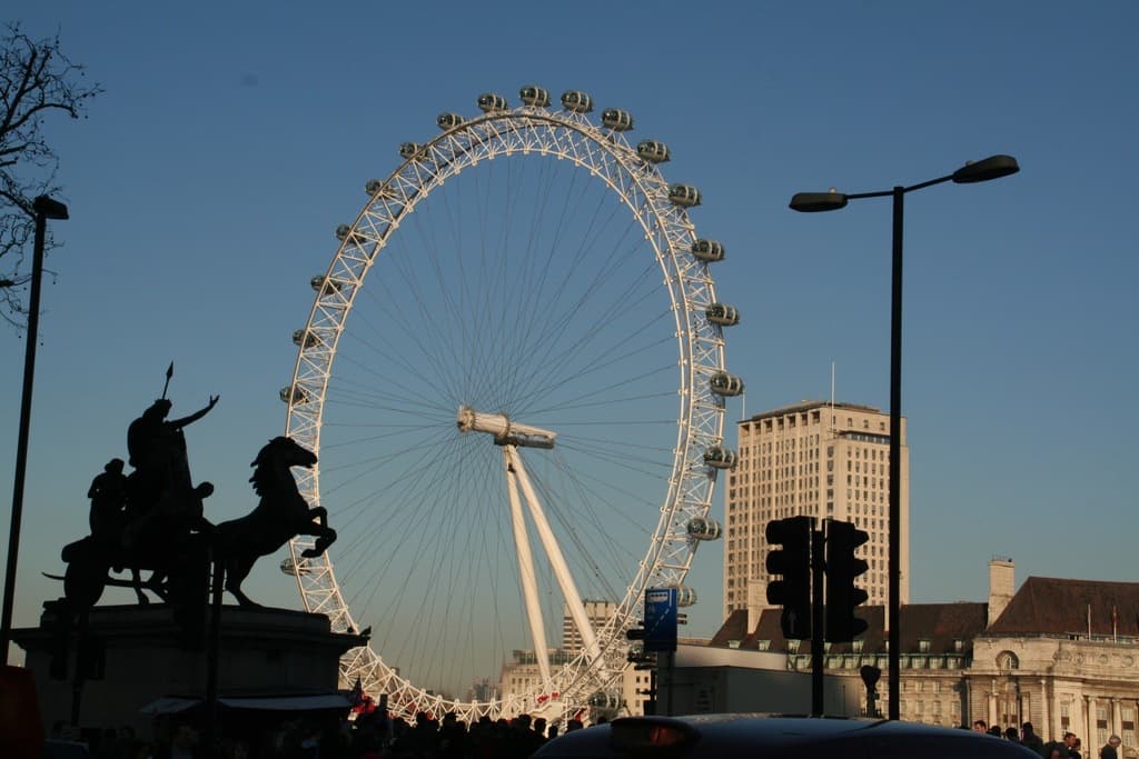 Gebäude und Pfähle auf der rechten Seite, ein großes Riesenrad in der Mitte und eine Skulptur auf der linken Seite.
