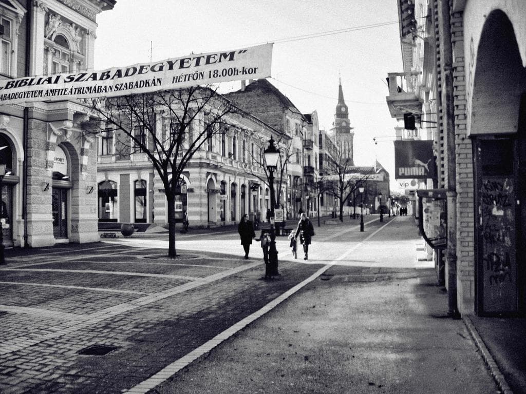 Straßenszene mit Gebäuden auf beiden Seiten, Banner oben links, Laubbäume, dekorative Laternen und einige Fußgänger