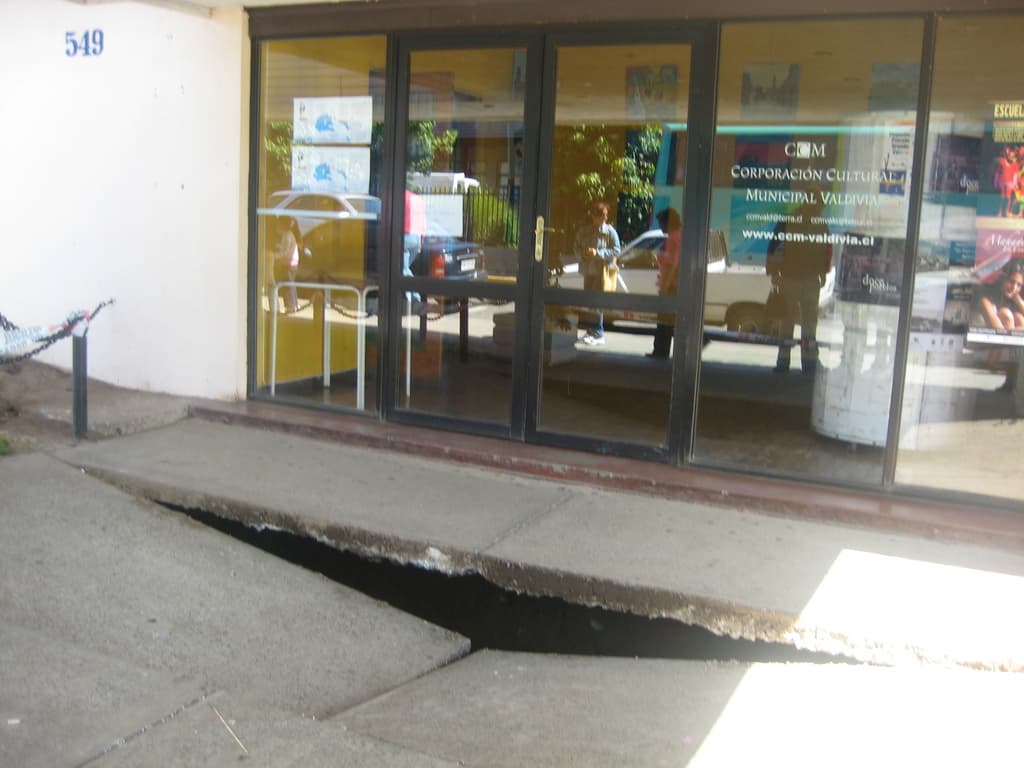 Eine gebrochene Oberfläche im Vordergrund mit einer Glastür, Wand und Pfahl im Hintergrund, die sich bewegende Fahrzeuge und einige stehende Personen spiegeln.