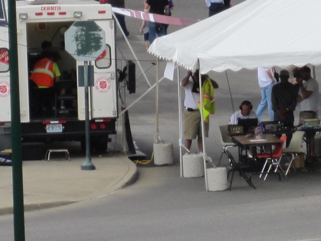 Ein Krankenwagen befindet sich auf der linken Seite einer Straße, während auf der rechten Seite ein Zelt eine Gruppe von Menschen schützt, mit einem Tisch und einem Stuhl in der Nähe.