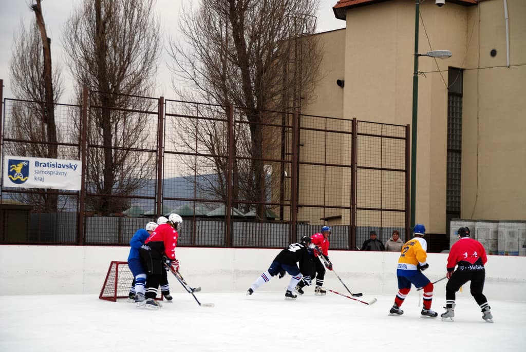 Menschen beim Eishockey-Spiel auf einer Eisfläche mit Gebäuden, Bäumen, einer Straßenlaterne, einem Namensschild und Zäunen im Hintergrund unter dem Himmel.