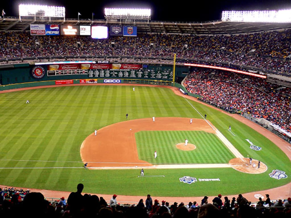 A baseball game in progress with players on the field, surrounded by spectators, hoardings, lights, and flags.