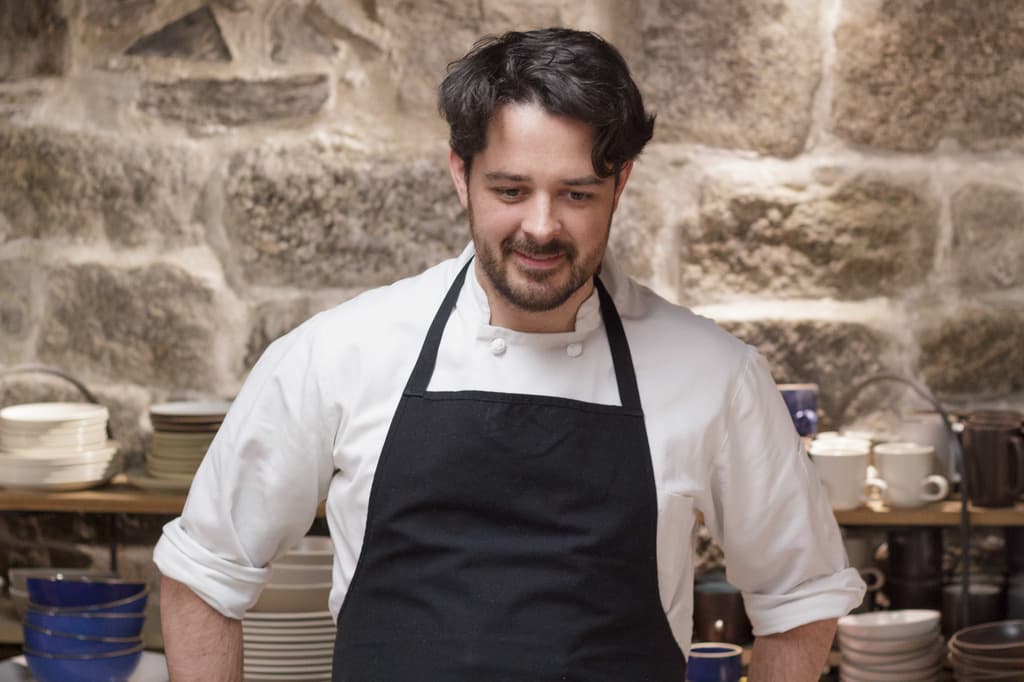 Ein Koch in weißem Hemd und schwarzer Schürze steht vor einem Regal mit Tellern, Schüsseln und Tassen an einer Wand.