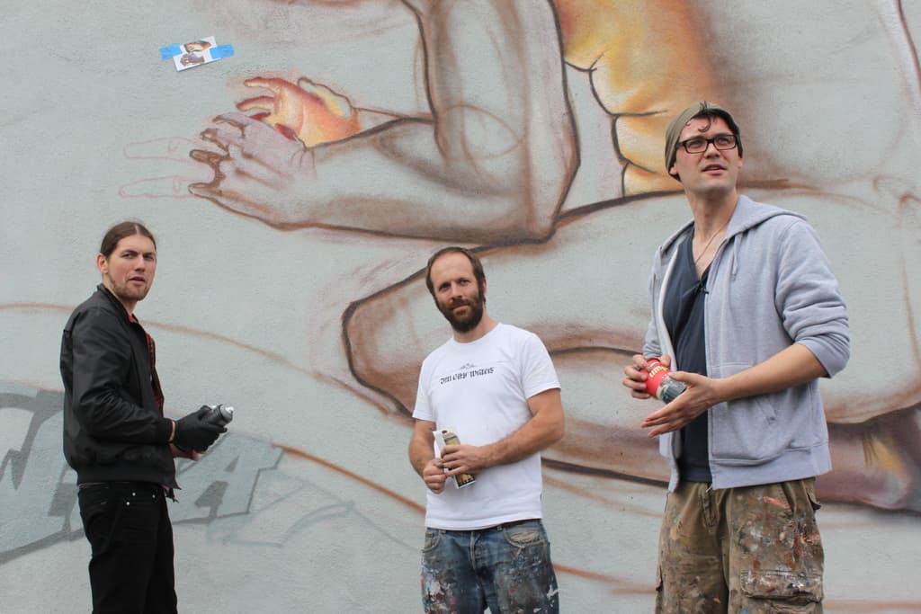 Menschen mit Farbsprühdosen in der Hand, mit Graffiti an der Wand dahinter.