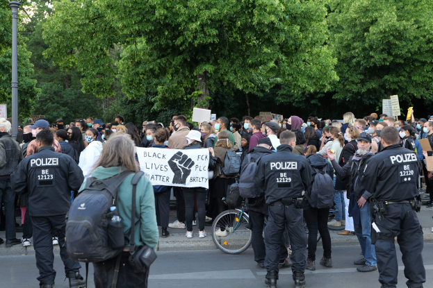 Eine große Gruppe von Menschen nimmt an einer Black-Lives-Matter-Demonstration in Berlin teil, einige halten Schilder, andere tragen Mützen und Taschen, mit einem Fahrrad im Vordergrund und Bäumen und einem Pfahl im Hintergrund.