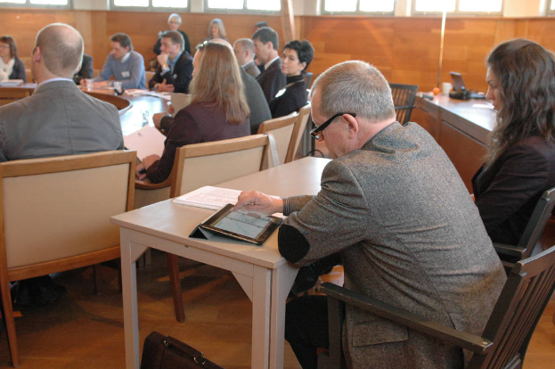 Zwei Personen sitzen auf schwarzen Stühlen in einem Büroraum, eine hält ein Tablet und die andere ein Buch, mit mehreren anderen Personen um einen runden Tisch mit Gläsern, Wasser und anderen Gegenständen, alle tragen ID-Karten, und ein Telefon auf einem Tisch rechts.