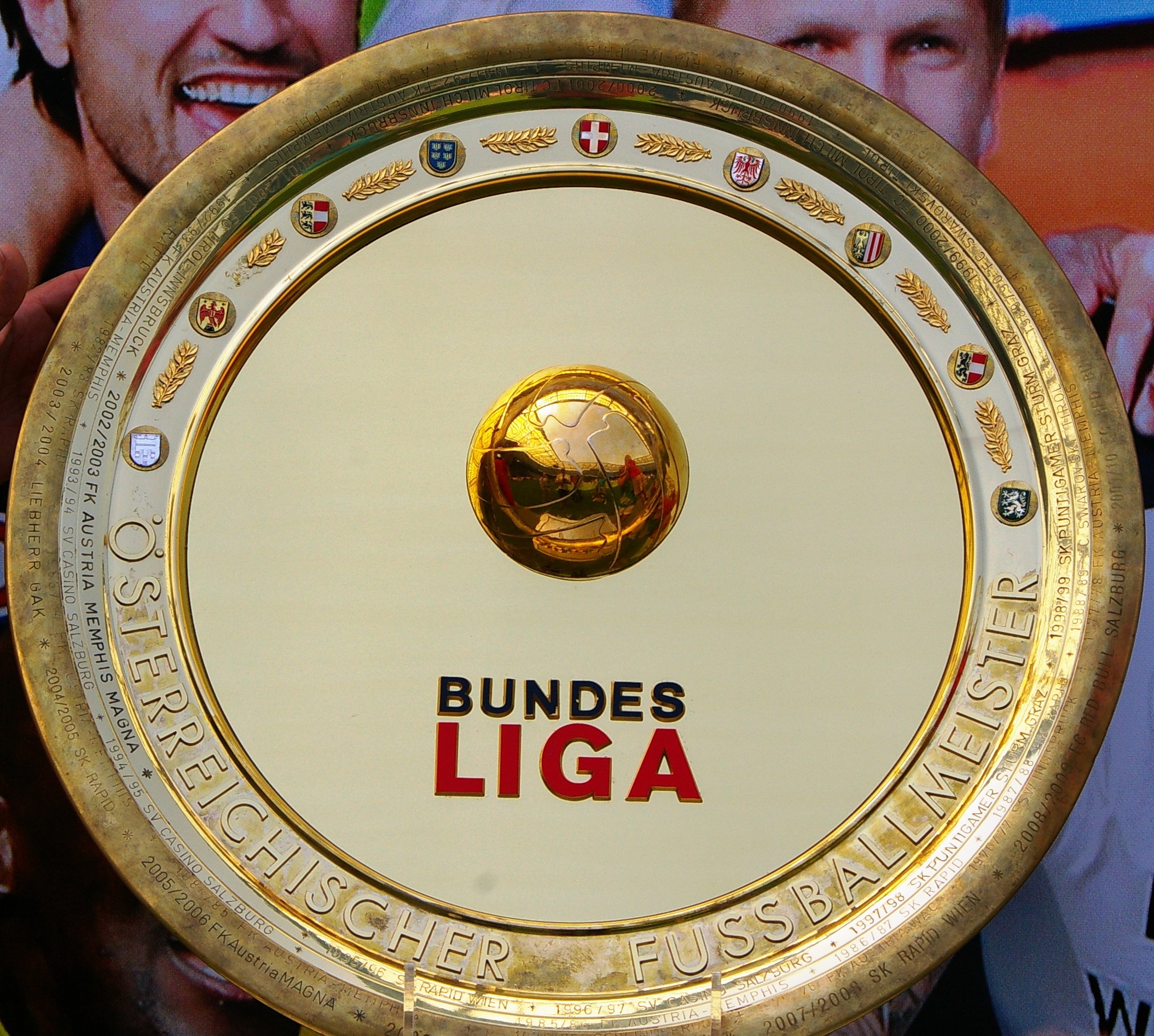 Ein Mann mit einem goldenen Schild mit der Aufschrift "Bundesliga", im Hintergrund eine Fahne mit drei Personen.