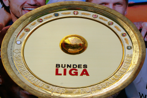 Ein Mann mit einem goldenen Schild mit der Aufschrift "Bundesliga", im Hintergrund eine Fahne mit drei Personen.