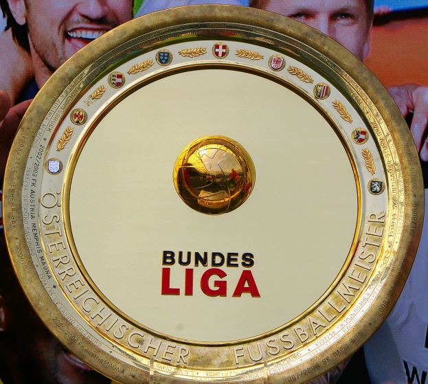 Ein Mann mit einem goldenen Schild mit der Aufschrift "Bundesliga", im Hintergrund eine Fahne mit drei Personen.
