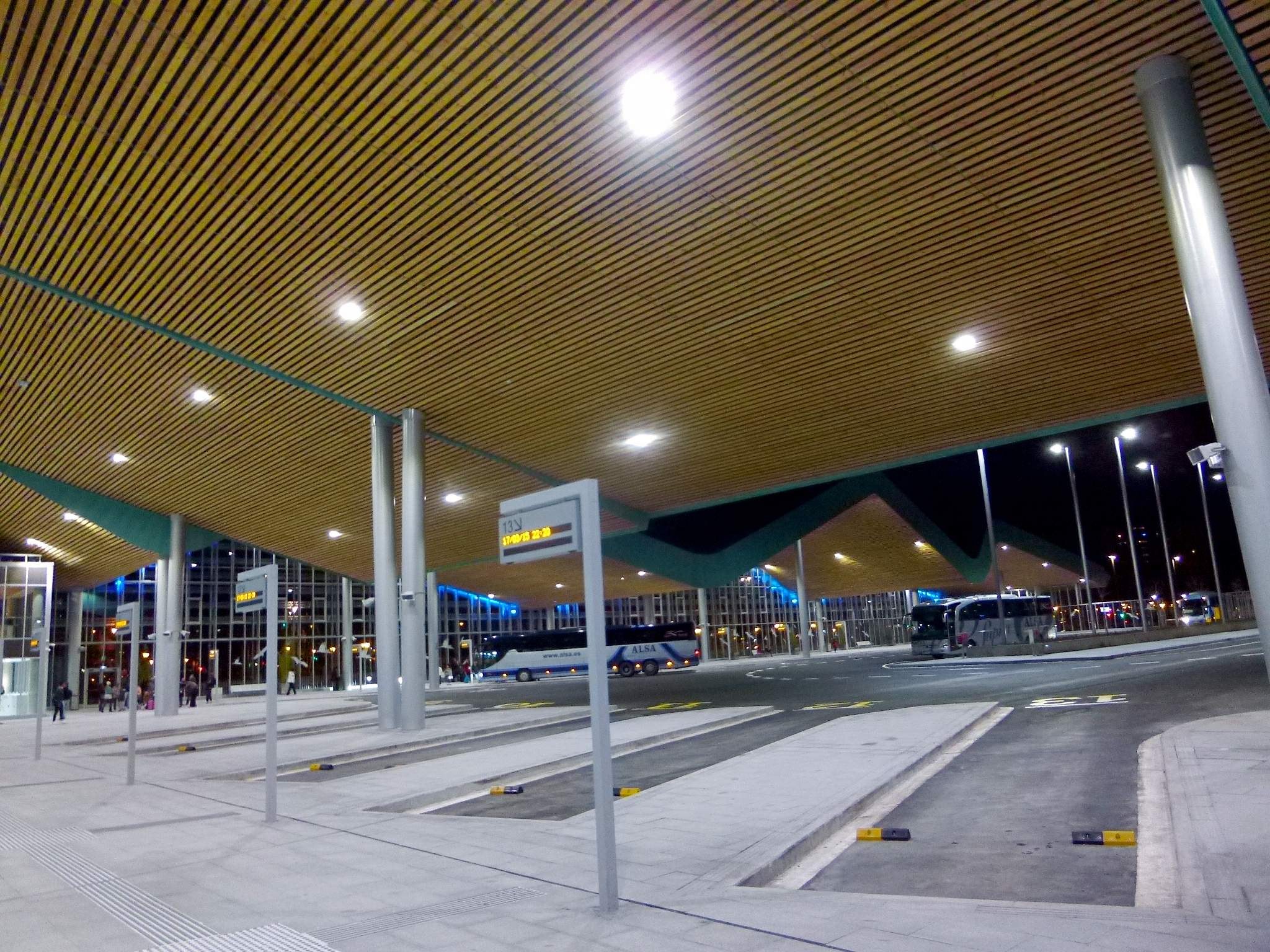 Eine Bushaltestelle nachts mit Deckenbeleuchtung, Fahrzeugen auf der Straße, Pfählen, Texttafeln, einigen Menschen und beleuchteten Gebäuden im Hintergrund.