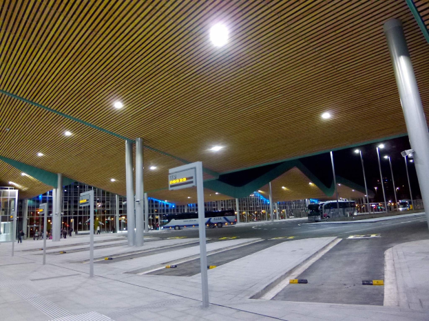 Eine Bushaltestelle nachts mit Deckenbeleuchtung, Fahrzeugen auf der Straße, Pfählen, Texttafeln, einigen Menschen und beleuchteten Gebäuden im Hintergrund.