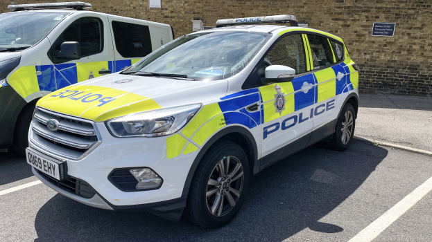Zwei Polizeiwagen nebeneinander auf einem Parkplatz geparkt, mit einer Wandtafel im Hintergrund, die Text zu einer Polizeiermittlung anzeigt.