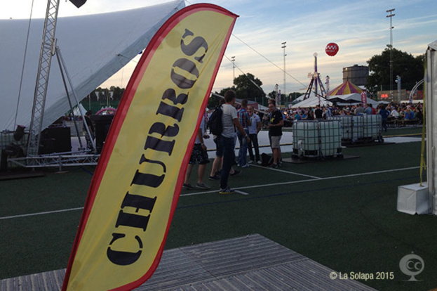 Ein lebhaftes Karnevalsszenario mit Zelten, einem zentralen Mast, Menschen, Bäumen, Strommästen, Straßenbahnen und einem eisernen Ständer auf der linken Seite.