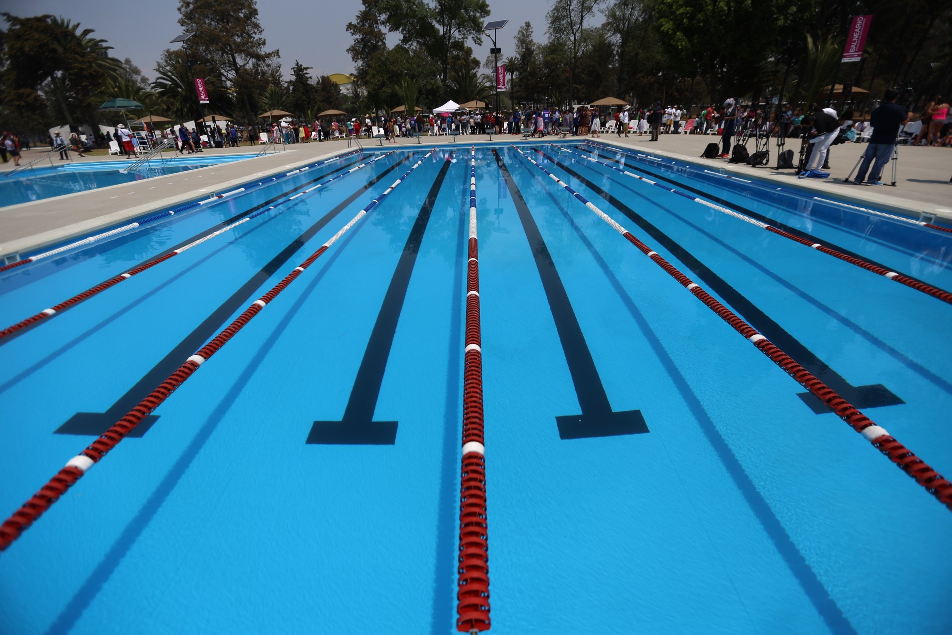 Großer Schwimmbad mit Bahnentrennung, umgeben von Menschen mit Taschen und Gegenständen, Zelte, Banner, Stangen, ein Schild und einen bewölkten Himmel im Hintergrund.