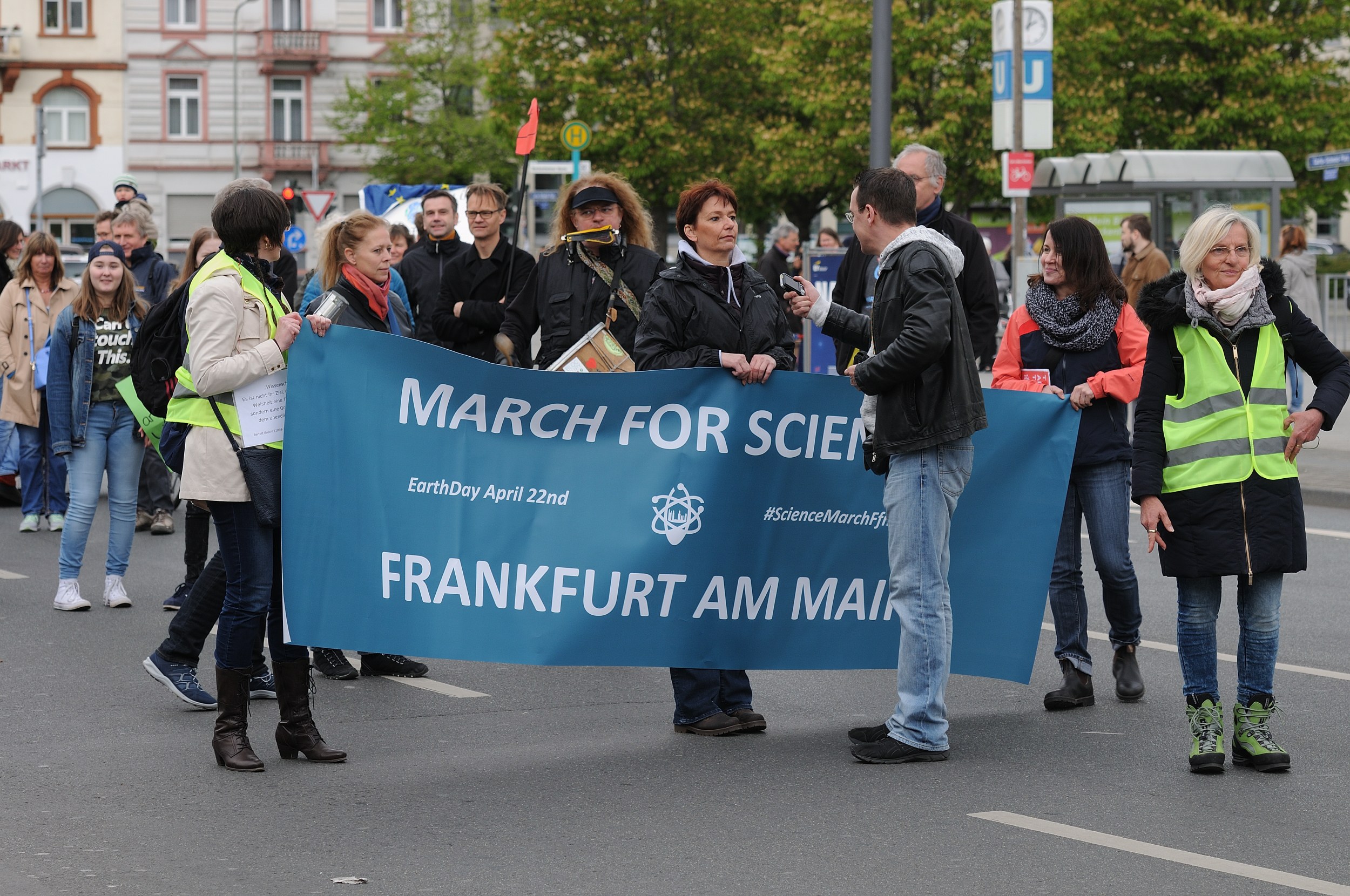 Gruppe von Menschen marschiert die Straße entlang und hält ein 'March for Science Frankfurt am Main'-Schild mit Bäumen, Pfählen, Schildern, Gebäuden und einem klaren blauen Himmel im Hintergrund.
