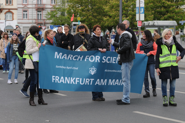 Gruppe von Menschen marschiert die Straße entlang und hält ein 'March for Science Frankfurt am Main'-Schild mit Bäumen, Pfählen, Schildern, Gebäuden und einem klaren blauen Himmel im Hintergrund.
