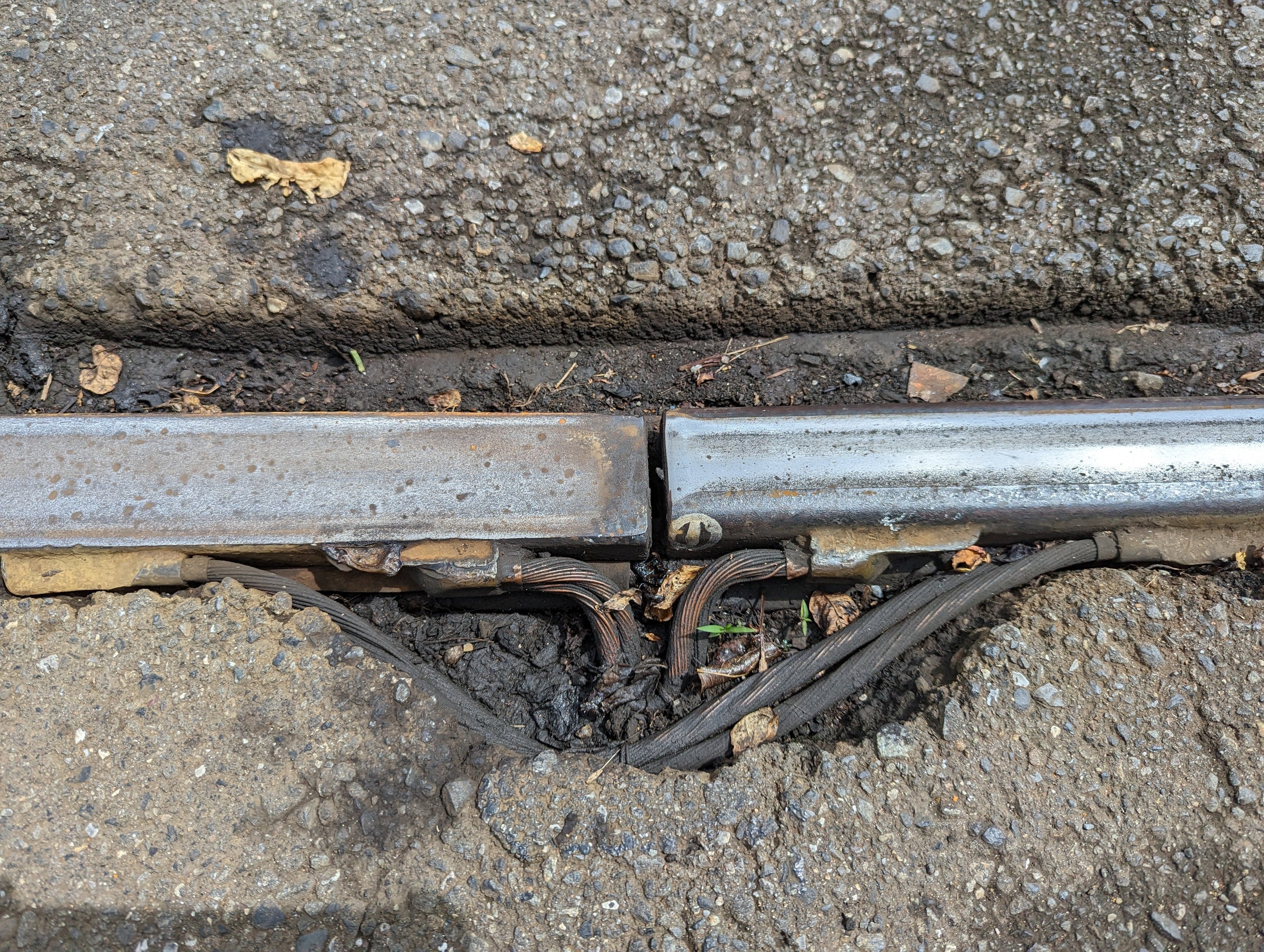 Nahaufnahme eines Metallkanalrohrs auf der Straße mit angebrachten Drähten und verstreuten trockenen Blättern.