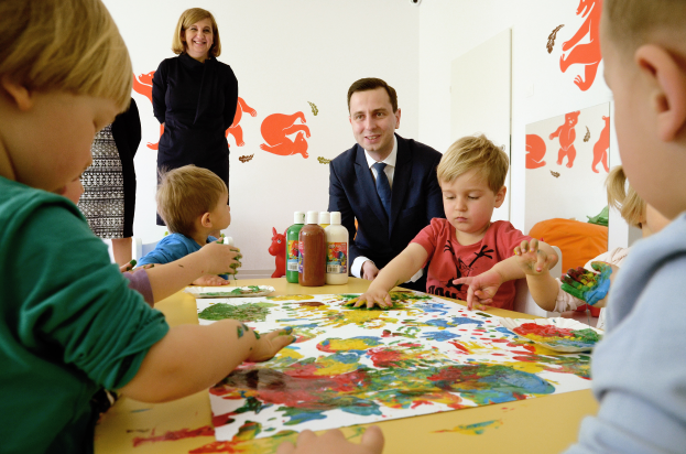 Eine Gruppe von Kindern mit Farbe an den Händen sitzt um einen Tisch mit Papieren und Flaschen, während eine lächelnde Frau danebensteht, mit Gemälden an der Wand im Hintergrund.