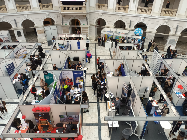 Große Besuchergruppe auf einer internationalen Fachmesse mit Ständen, Bannern, Tischen, Stühlen und Ballons in einem Raum mit Bogengängen, Säulen und einem Bildschirm im Hintergrund.