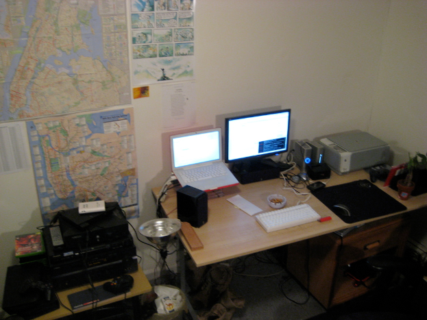 Ein Raum mit einem Tisch, auf dem ein Laptop, Lautsprecher, Monitor, Tastatur, Ball, Maus, Scanner und Papiere sowie ein weiterer Tisch mit Elektronikgeräten stehen, und Charts und Plakate an der Wand dahinter.