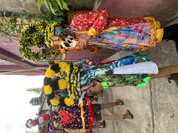 Eine Gruppe von Menschen in bunten Kostümen steht vor einem Gebäude und hält verschiedene Gegenstände, mit Bäumen, Gebäuden und einem klaren blauen Himmel im Hintergrund während eines Karnevalsumzugs.