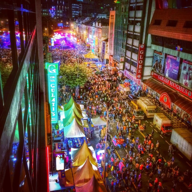 Ein lebhaftes Karneval-Szenario mit einer großen Menschenmenge auf der Straße, umgeben von Zelten, Geschäften, Gebäuden und Bannern, mit zahlreichen Lichtern im Hintergrund.
