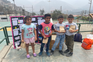 Eine Gruppe von Kindern steht auf einem Basketballfeld, jedes hält einen Papiertüte in der Hand, mit Tüten und Gegenständen rechts und einer Tafel mit Text links, umgeben von Zaun, Pfosten, Straßenlaternen, Bäumen, Gebäuden, Hügeln und dem Himmel.