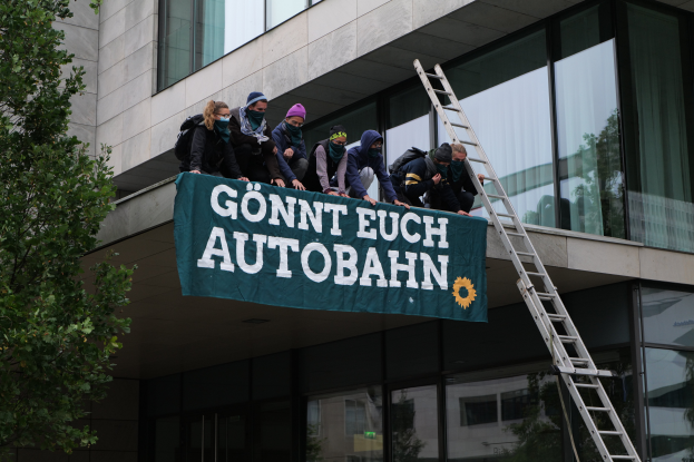 Eine Gruppe von Menschen steht auf dem Dach eines Gebäudes, hält eine Fahne, mit einem Baum links und Glasfenstern im Hintergrund, wahrscheinlich protestiert sie im Namen der deutschen Regierung.