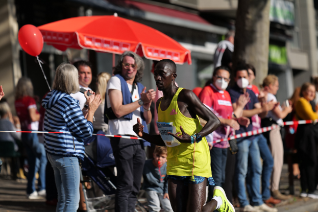 Ein Mann in einem gelben T-Shirt und Laufschuhen sprintet durch einen Marathon, während ihn eine Menge anfeuert und klatscht, mit einem Schirm und einem Kinderwagen im Hintergrund.