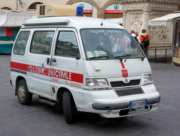 Ein weißer Van fährt auf einer Straße mit einer Gruppe von Menschen in der Nähe, einem Schuppen auf der rechten Seite und Gebäuden und Metallstangen im Hintergrund.