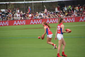 Zwei Frauen in Sportbekleidung beim Australian Football League-Spiel auf einem Feld, eine hält den Ball, mit Zuschauern und Werbetafeln im Hintergrund.