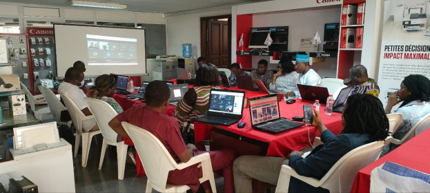 Eine Gruppe von Menschen sitzt um einen Tisch mit Laptops, Müsen und Flaschen und nimmt an einem digitalen Marketing-Workshop teil, mit einem Banner, einem Display-Bildschirm und elektronischen Geräten im Hintergrund.
