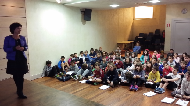 Eine Gruppe von Kindern, Jungen und Mädchen, sitzt auf dem Boden mit Büchern vor sich, während eine Frau auf der linken Seite auf einer Bühne steht.