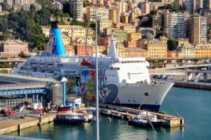 Ein großes Kreuzfahrtschiff liegt nahe einer Stadt mit einer Brücke links, Fahrzeugen auf der Straße und Gebäuden und Bäumen im Hintergrund.