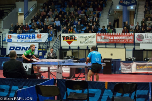 Indoor-Tischtennis-Match mit zwei Spielern und mehreren stehenden Zuschauern, sitzendem Publikum, leeren Stühlen und Werbetafeln in einer Stadionsetting.