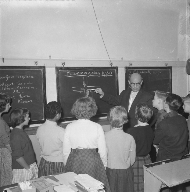 Ein Schwarz-Weiß-Foto eines Lehrers, der vor einer Tafel steht und einen Marker h"alt, umgeben von Kindern an einem Tisch mit B"chern und anderen Gegenst"anden, und einem Lautsprecher an der Wand rechts.