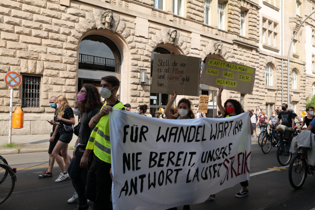 Eine Gruppe von Menschen marschiert auf einer Stadtstraße bei einer Protestaktion, einige halten Schilder und andere fahren Fahrräder, im Hintergrund ein historisches Gebäude mit Rundbogenfenstern, Säulen und Skulpturen.