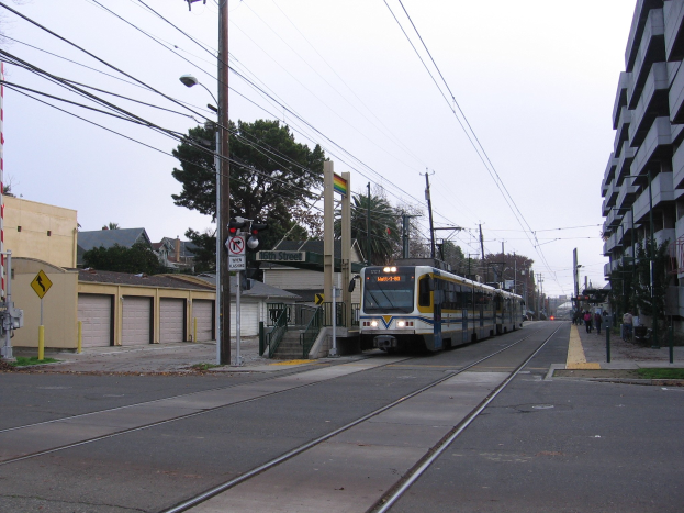 Eine Stadtbahn fährt auf einer Straße mit hohen Gebäuden, Strommasten mit Drähten, Fußgängern auf dem Gehweg, Bäumen im Hintergrund und einem klaren blauen Himmel.