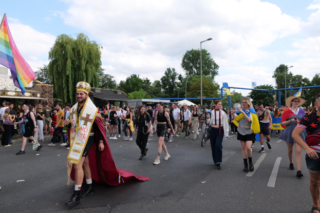 Eine Gruppe von Menschen marschiert bei der Gay Pride Parade 2018 mit einer Regenbogenflagge und Musikinstrumenten, umgeben von Laternenmasten, Bäumen, Schuppen und einem bewölkten Himmel.