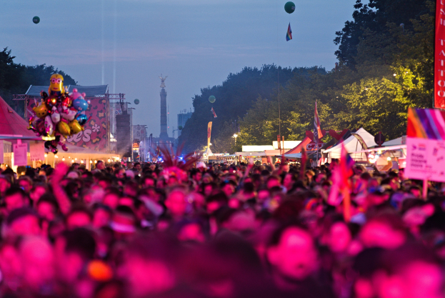 Eine große Menschenmenge auf einem nächtlichen Musikfestival mit farbigen Lichtern, Ballons, Zelten, Bannern, Fahnen, Bäumen, einem Turm und einem sternenklaren Himmel im Hintergrund.