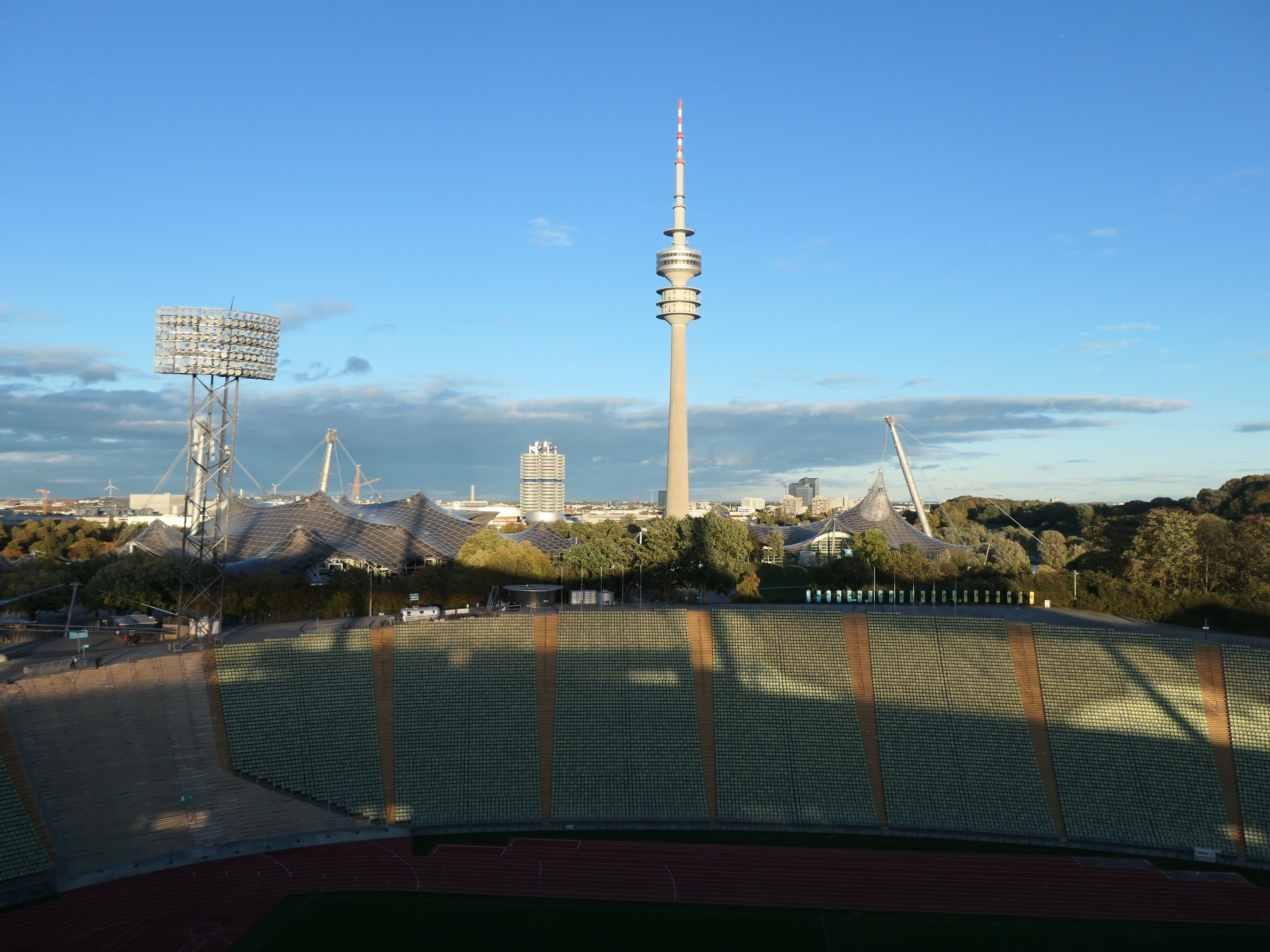 Das Olympiastadion in Berlin, Deutschland, mit dem Fernsehturm (Fernsehturm) im Hintergrund, umgeben von Bäumen, Gebäuden und beleuchteten Strukturen unter einem bewölkten Himmel.