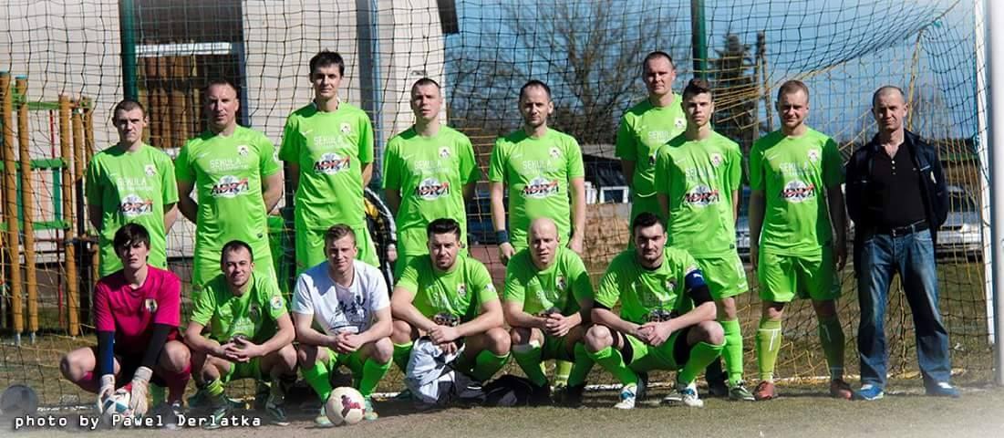 Mannergruppe in grünen T-Shirts auf einem Fußballfeld mit einem Ball, einige in Hockstellung, neben einem Torpfosten, mit Bäumen, Gebäuden und Fahrzeugen im Hintergrund.