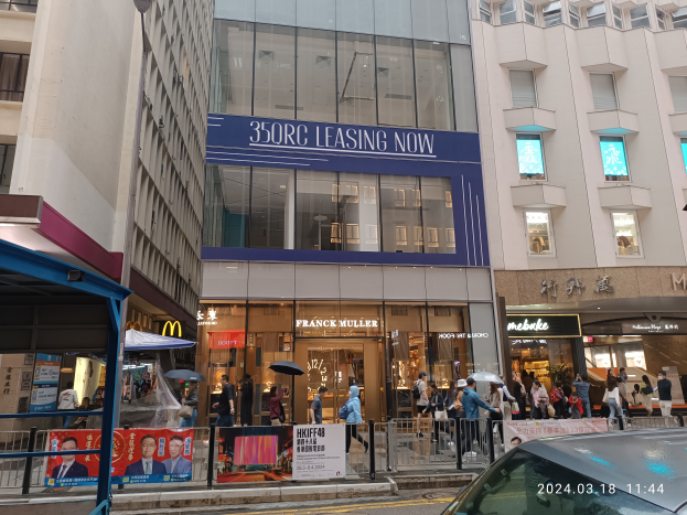 Eine belebte Stadtstraße mit einem geparkten Auto vor einem Gebäude mit einer '350rc Leasing Now'-Schilder, Menschen mit Regenschirmen, Geländern, Bannern und einer Laterne im Hintergrund.