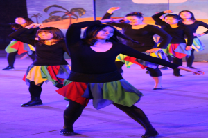 Gruppe junger Frauen in bunten Röcken und Schuhen, die energisch auf einer Bühne tanzen, mit einem Gemälde an der Wand im Hintergrund.