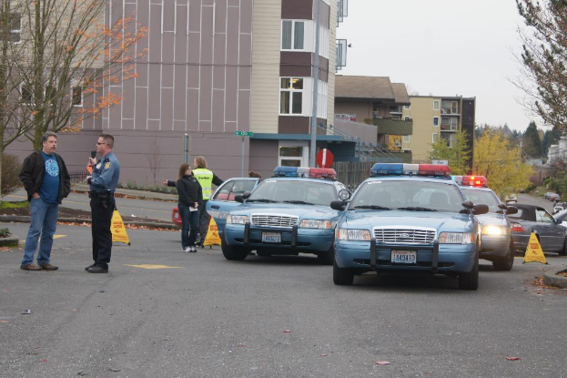 Autos auf einer StraÃŸe mit vier Menschen in der NÃ¤he, GebÃ¤ude mit Fenstern im Hintergrund, BÃ¤ume und Warnkegel.