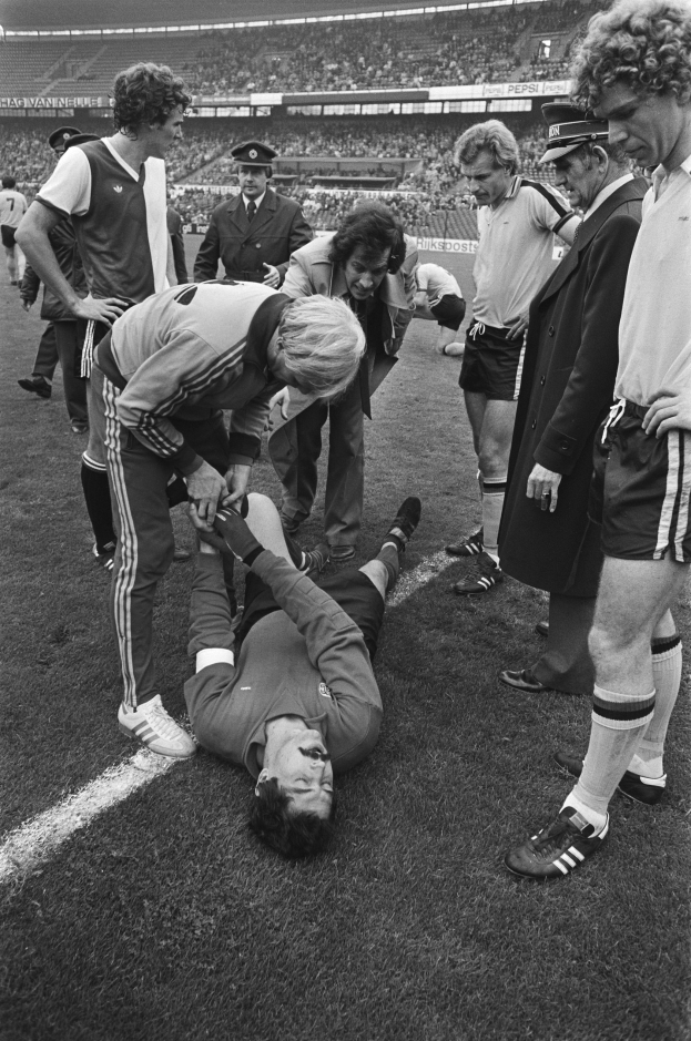 Schwarzes und weißes Bild eines Fußballspielers, der auf dem Boden liegt und von einer Gruppe von Männern umgeben ist, mit einer Zuschauermenge im Hintergrund eines Stadions.