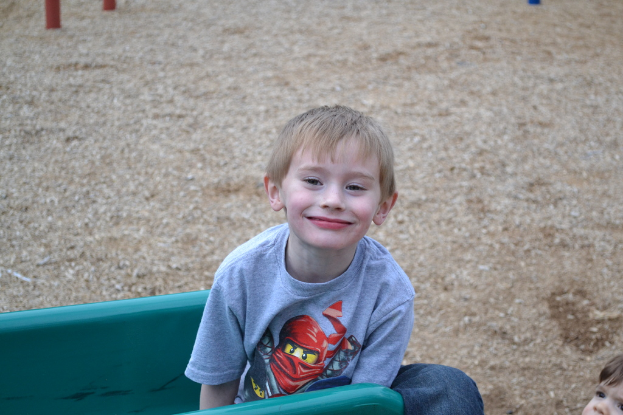 Ein Kind auf einer Bank sitzend, das lächelt, mit einem anderen Kind auf der rechten Seite und Pfählen oben im Bild.