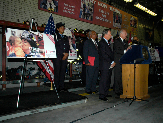 Eine Gruppe von Menschen in der Nähe eines Podiums mit Mikrofonen, Schildern auf Ständern, Flaggen und Schildern an der Wand, alles unter hellem Licht.