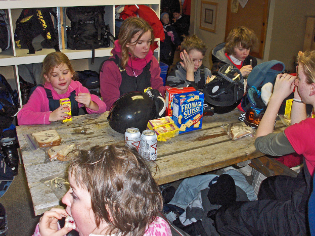 Eine Gruppe von Kindern sitzt um einen Esstisch und isst Essen, mit Taschen an einem Gestell oben und Lebensmitteln auf dem Tisch.